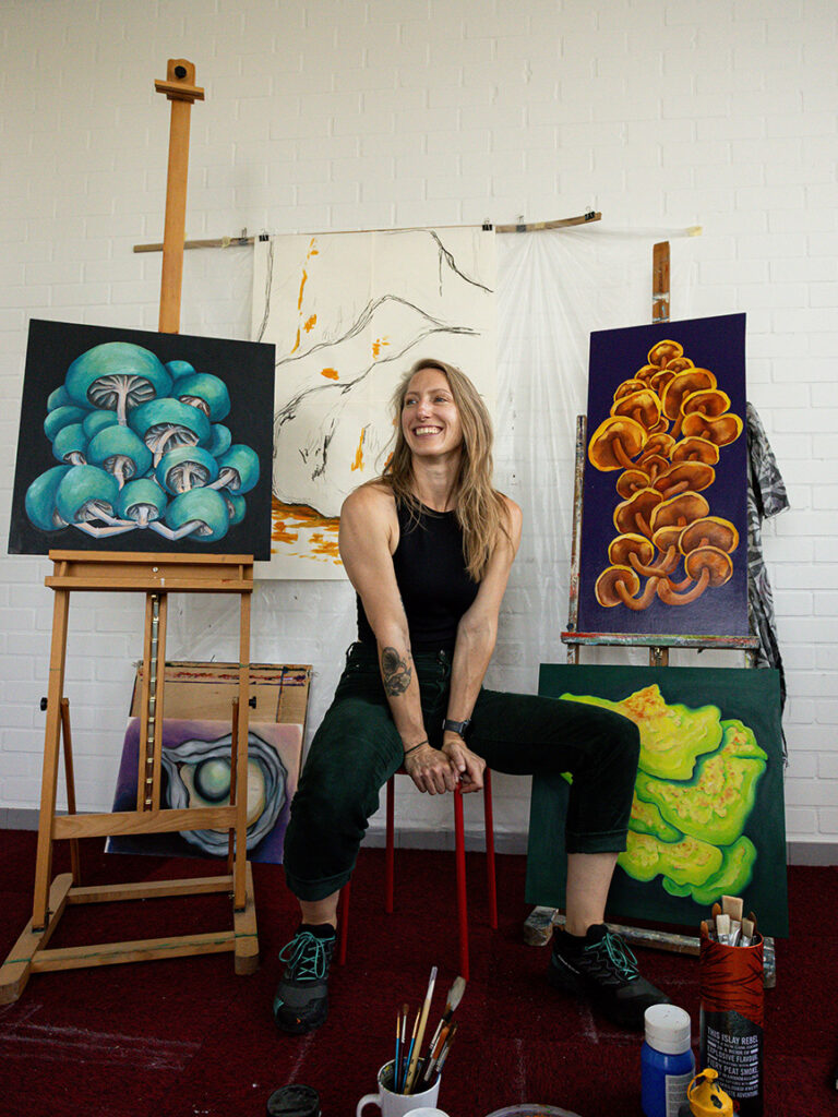 portret kunstenaar, zittend, omringd door schilderijen van kleurrijke paddenstoelen